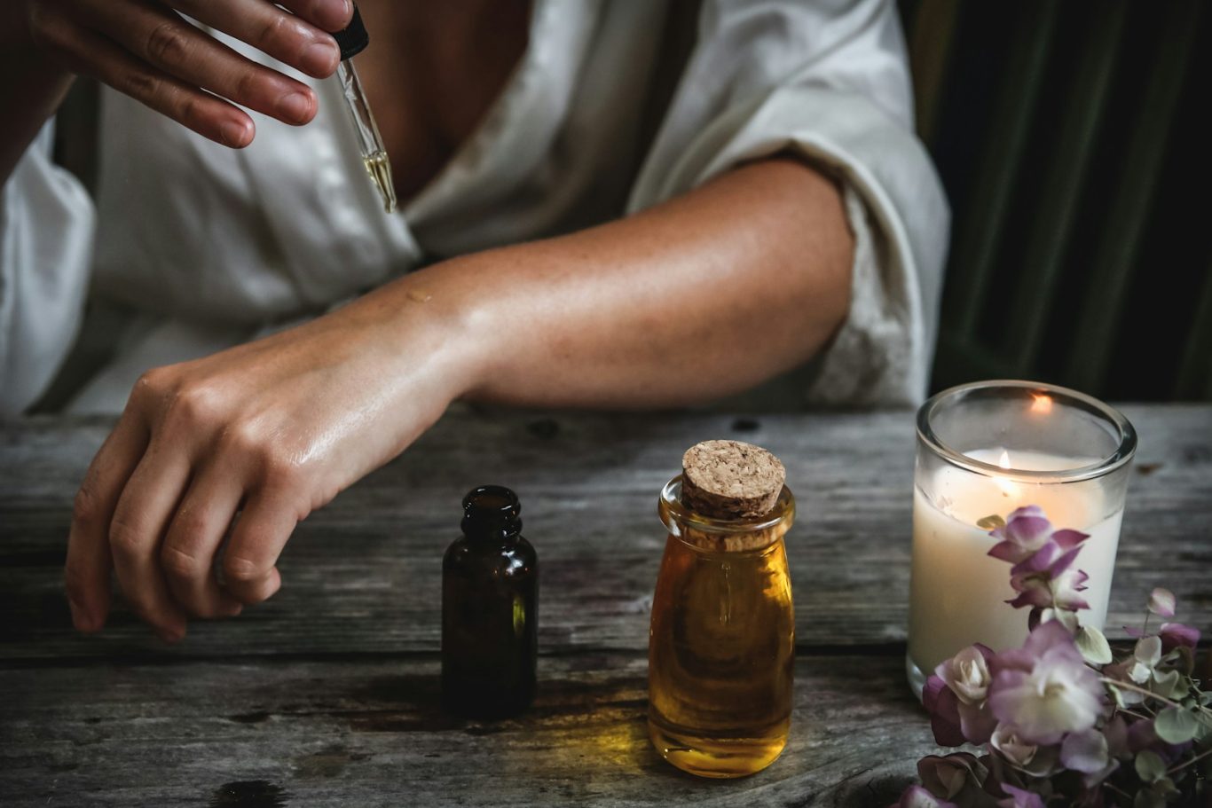A hand applying essential oil next to a lit candle and a bottle of oil on a wooden surface.