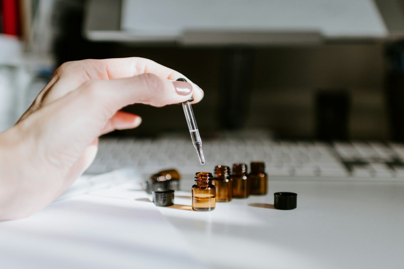 A hand holding a dropper above several small amber glass vials on a white surface.